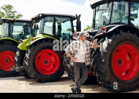 Händler oder Lieferant von Landmaschinen. Kaukasischer Mann im karierten Hemd steht neben Traktoren und benutzt ein digitales Tablet. Stockfoto
