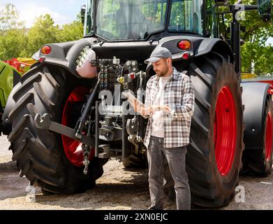 Händler oder Lieferant von Landmaschinen. Kaukasischer Mann in kariertem Hemd steht neben dem Traktor und benutzt ein digitales Tablet. Stockfoto