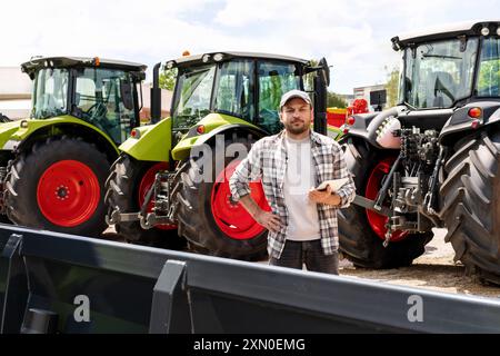 Kaukasischer Erwachsener mit digitalem Tablet in der Hand steht neben einem Bau- oder landwirtschaftlichen Traktor bei einem Maschinenhändler. AG-Gerätehändler oder -Zulieferer Stockfoto