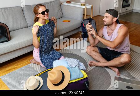 Ein Paar sitzt zu Hause auf dem Boden, packt oder auspackt Reisetaschen und probiert ein Kleid an, während ein Mann eine Frau fotografiert, während sie ein Kleid anprobiert. Stockfoto