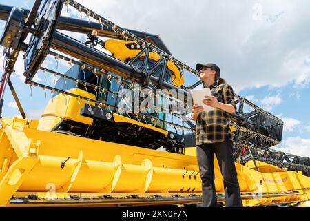 Händler oder Lieferant von Landmaschinen. Frau im karierten Hemd hält ein digitales Tablet in der Hand und sieht den gelben Mähdrescher an. Stockfoto