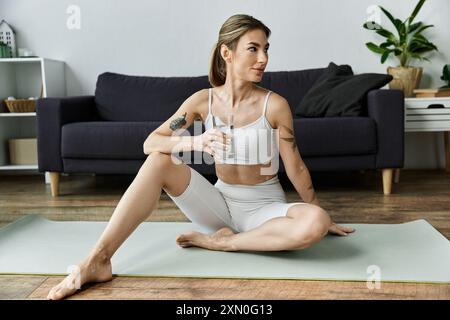 Eine junge Frau in Activewear sitzt auf einer Yogamatte in einem modernen Apartment und hält nach einer Yoga-Session ein Glas Wasser in der Hand. Stockfoto