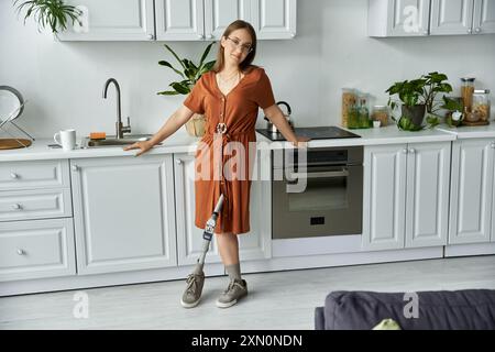 Eine Frau in braunem Kleid lächelt selbstbewusst in ihrer Küche, ihre Beinprothese ist sichtbar. Stockfoto