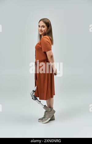 Eine Frau mit einer Beinprothese steht selbstbewusst in einem braunen Kleid vor weißem Hintergrund. Stockfoto