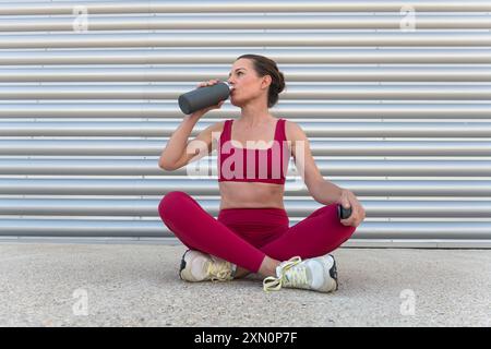 Sportliche Frau, die nach dem Training im Freien ausruht und Wasser aus einer Flasche trinkt, mit überkreuzten Beinen sitzend Stockfoto