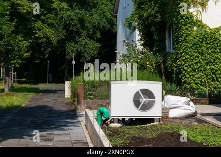 Ein Techniker arbeitet an der Installation einer Klimaanlage in einem üppigen Wohngarten, umgeben von Grün an einem hellen, sonnigen Nachmittag. Stockfoto