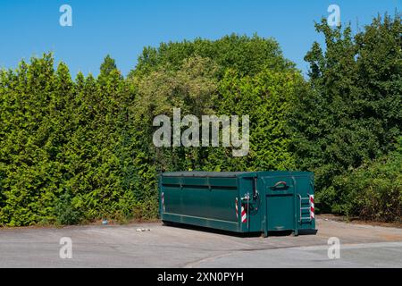 Ein grüner Abfallbehälter steht ruhig in einem städtischen Gebiet, eingerahmt von hohen, dichten Bäumen unter einem klaren blauen Himmel. Stockfoto