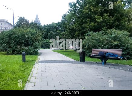 Moskau, Russland, August 2019: Ein gut gekleideter junger Mann schläft auf einer Bank in einem sehr grünen Sommerpark. Obdachlos oder betrunken. In der Nähe - eine Flasche Wasser. Stockfoto