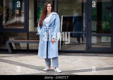 In Übergröße steht eine junge Frau im Freien, modernes urbanes Ambiente, zeitgenössische Stadt, mit stilvollem hellblauem Trenchcoat, weißem T-Shirt und blauer Jeans Stockfoto