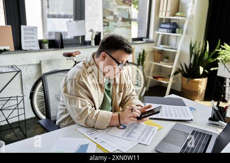 Ein junger Mann mit Down-Syndrom sitzt an seinem Schreibtisch und konzentriert sich auf sein Handy. Stockfoto