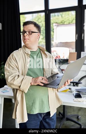 Ein junger Mann mit Down-Syndrom, Brille trägt, steht mit einem Laptop in einem Büro. Stockfoto