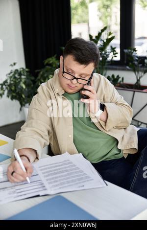 Ein junger Mann mit Down-Syndrom sitzt an einem Schreibtisch und macht sich Notizen während eines Telefonats. Stockfoto