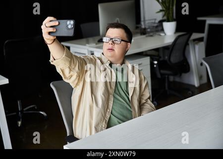 Ein junger Mann mit Down-Syndrom sitzt an einem Schreibtisch und macht ein Selfie mit seinem Handy. Stockfoto