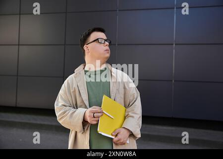 Ein Mann mit Down-Syndrom, Brille, beigefarbener Jacke steht draußen und hält einen gelben Ordner Stockfoto