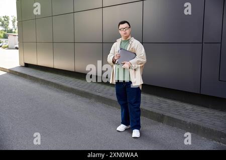 Ein junger Mann mit Down-Syndrom steht vor einem modernen Gebäude und hält einen Laptop in der Hand. Stockfoto