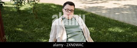 Ein junger Mann mit Down-Syndrom sitzt auf einer Bank im Park, trägt Brille und eine leichte Jacke. Stockfoto