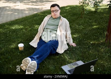 Ein junger Mann mit Down-Syndrom sitzt auf dem Gras mit einer Kaffeetasse und einem Laptop. Stockfoto