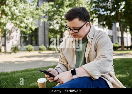 Ein junger Mann mit Down-Syndrom sitzt auf einer grasbewachsenen Fläche in einem Park und benutzt sein Smartphone. Stockfoto