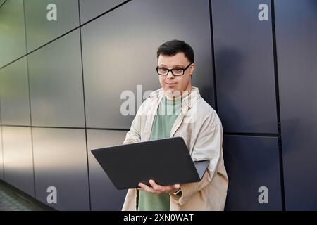 Ein junger Mann mit Down-Syndrom steht draußen und lächelt, während er einen Laptop benutzt. Stockfoto