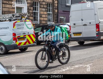 Edinburgh, Schottland, 07.17.2024: Fahrer mit Essen auf einem Elektrofahrrad lächelnd, während er die Straße entlang radelte, Kopfhörer, Helm, Deliveroo j. Trug Stockfoto