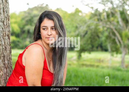 lateinische Frau, die zur Seite schaut, sich an einen Baum mit grünem Hintergrund lehnt. Stockfoto