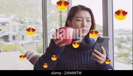 Bild von Emoji-Symbolen über asiatische Frau, die Kaffee trinkt und ihr Smartphone zu Hause benutzt Stockfoto