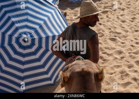 Tanger, Marokko. Juli 2024. Ein Mann führt ein Kamel zwischen den Sonnenschirmen an einem Strand in Tanger in Marokko. (Credit Image: © David Canales/SOPA Images via ZUMA Press Wire) NUR REDAKTIONELLE VERWENDUNG! Nicht für kommerzielle ZWECKE! Stockfoto