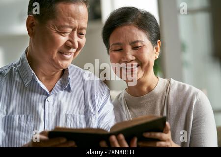 Glückliches älteres asiatisches Paar, das zu Hause auf der Familiencouch sitzt und zusammen Bibel liest Stockfoto