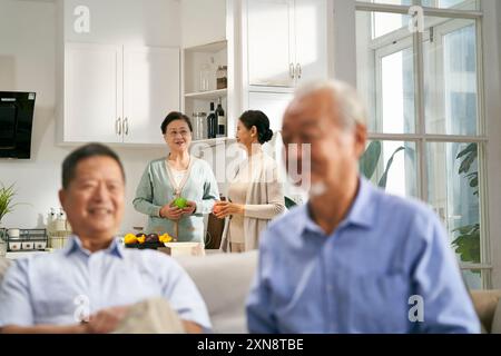 Zwei glückliche ältere asiatische Frauen sitzen und plaudern zu Hause auf der Familiencouch mit Ehemännern im Vordergrund Stockfoto