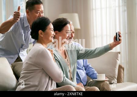 Gruppe glücklicher älterer asiatischer Leute zwei Paare, die zu Hause auf der Familiencouch sitzen und gemeinsam ein Selfie mit dem Handy machen Stockfoto