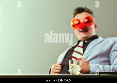 Ein Geschäftsmann sitzt an seinem Schreibtisch mit einem Glas Whiskey vor ihm, trägt eine Clownnase und eine Brille. Er scheint sich zu amüsieren Stockfoto