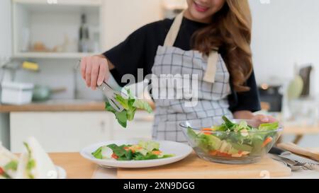 Nahaufnahme Hände der jungen asiatischen Frau mit Zange, um Salat in den Teller zu nehmen, nachdem frischen Gemüse Salat in der Schüssel gemischt und gesundes Frühstück gekocht Stockfoto