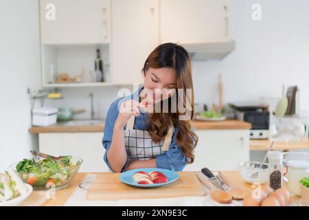 Junge asiatische Frau genießt und isst Scheiben Apfel während der Zubereitung von Obst und frischem Gemüse Salat zu kochen gesundes Frühstück Essen für hausgemachte sn Stockfoto