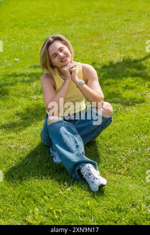 Eine junge Erwachsene Frau lächelt hell, während sie einen sonnigen Tag auf grünem Gras im Park genießt. Ihre lässige Kleidung und ihr fröhliches Auftreten spiegeln eine Pflege wider Stockfoto