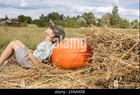 An einem sonnigen Tag liegt großer Kürbis auf trockenem Gras, der Junge lehnt sich träumerisch darauf und entspannt in der Natur. Das Kind hilft im Herbst, Gemüse auf dem Bauernhof zu sammeln Stockfoto