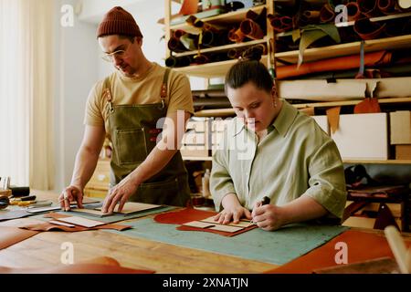 Der Schneider und seine Kollegin mit Down-Syndrom schneiden Lederdetails in einem mit Nähwerkzeugen und Stoffen ausgestatteten, antiken Atelier aus Stockfoto