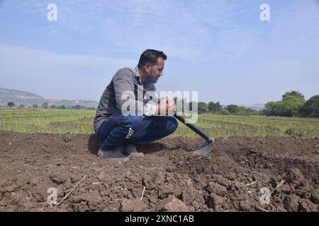 Junger Landwirt, der mit einer Schaufel auf dem Feld sitzt, Hintergrund von weißem bewölktem Himmel und grünen Feldern Stockfoto