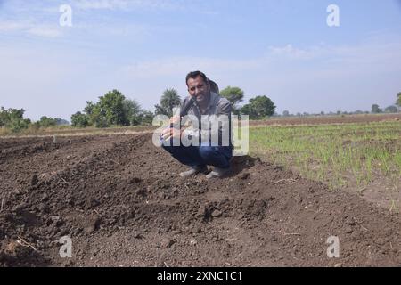Junger Landwirt, der mit einer Schaufel auf dem Feld sitzt, Hintergrund von weißem bewölktem Himmel und grünen Feldern Stockfoto