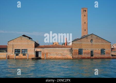 Salviati Murano Fabrikwände mit Blick auf das Wasser, die Spitze einer Kirche und ein hoher, kippbarer Glockenturm, blauer Himmel, türkisfarbenes Wasser in Murano, Venedig Italien Stockfoto