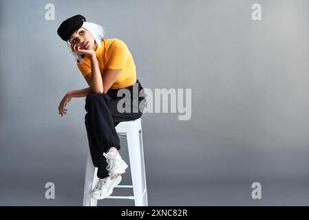 Frau, Stuhl und Mode im Studio mit Hut für Selbstvertrauen, Denken und Stilideen im Modell. Model Girl, Hocker und Streetwear-Kleidung mit Zeitungsjungen Stockfoto