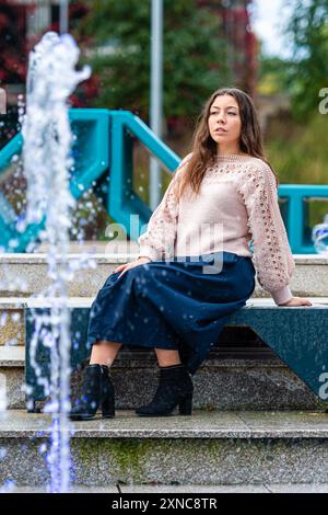 Eine junge Frau sitzt anmutig neben einem Brunnen in einem Stadtpark. Stockfoto