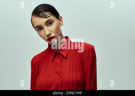 Eine schöne junge Frau mit kurzen Haaren, gekleidet in einem leuchtend roten Hemd, posiert selbstbewusst vor grauem Hintergrund. Stockfoto