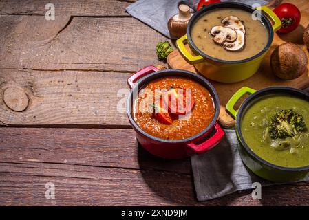 Herbstgemüsecremesuppen Set. Drei Portionstöpfe mit verschiedenen veganen heißen Gemüsesuppen – Tomaten, Pilze, Brokkoli auf rustikalem Holztisch Stockfoto