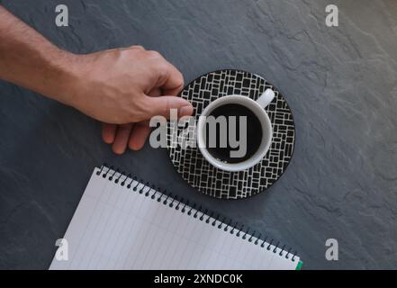 Hand eines weißen Mannes, der eine Tasse Kaffee neben einem kleinen Notizbuch mit einem grauen Schreibtisch im Hintergrund hält. Stockfoto