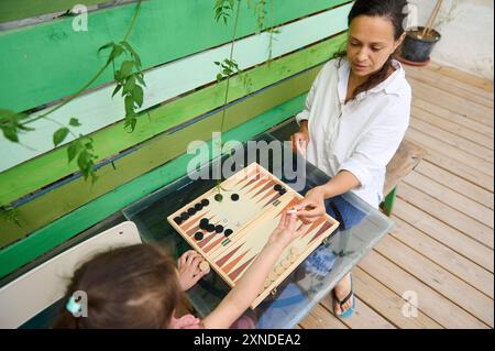 Mutter und Tochter spielen gerne gemeinsam Backgammon im Freien auf einer sonnigen Holzterrasse. Familienbeziehungen und Freizeit. Stockfoto