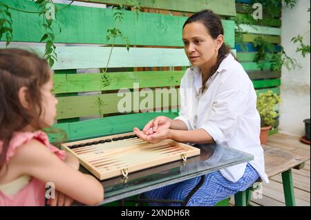 Eine Mutter und Tochter verbringen gute Zeit miteinander und spielen Backgammon auf einem Glastisch in ihrem Garten. Die Umgebung verfügt über grüne Holzzäune und Stockfoto