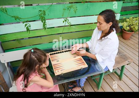 Mutter und Tochter spielen Backgammon draußen an einem sonnigen Tag. Familienverbundenheit und hochwertige Zeit, die Sie mit einem klassischen Brettspiel in einer Gartenanlage verbracht haben. Stockfoto