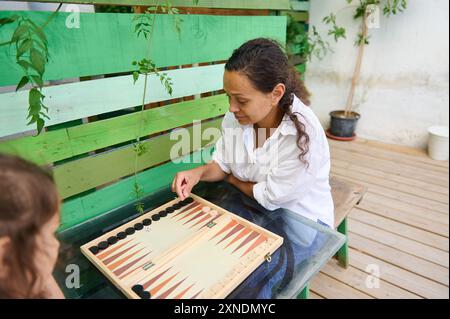 Eine Frau und ein Kind spielen auf einer hölzernen Terrasse im Freien mit grünem Hintergrund eine Runde Backgammon und genießen die Freizeitaktivität. Stockfoto