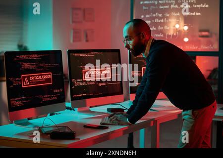 Seitenansicht Porträt eines erwachsenen Cybersicherheitsexperten, der am Notfall arbeitet und Computer verwendet, mit kritischer Fehlermeldung auf dem Bildschirm Stockfoto