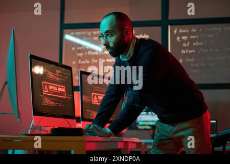 Dramatisches Porträt des bärtigen Cybersicherheitsexperten, der einen Notfall verhindert und Computer benutzt, mit roter kritischer Fehlermeldung Stockfoto